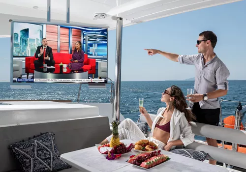 Couple enjoys drinks and snacks on a yacht, watching with the outdoor TV showing a news program in Kuwait.